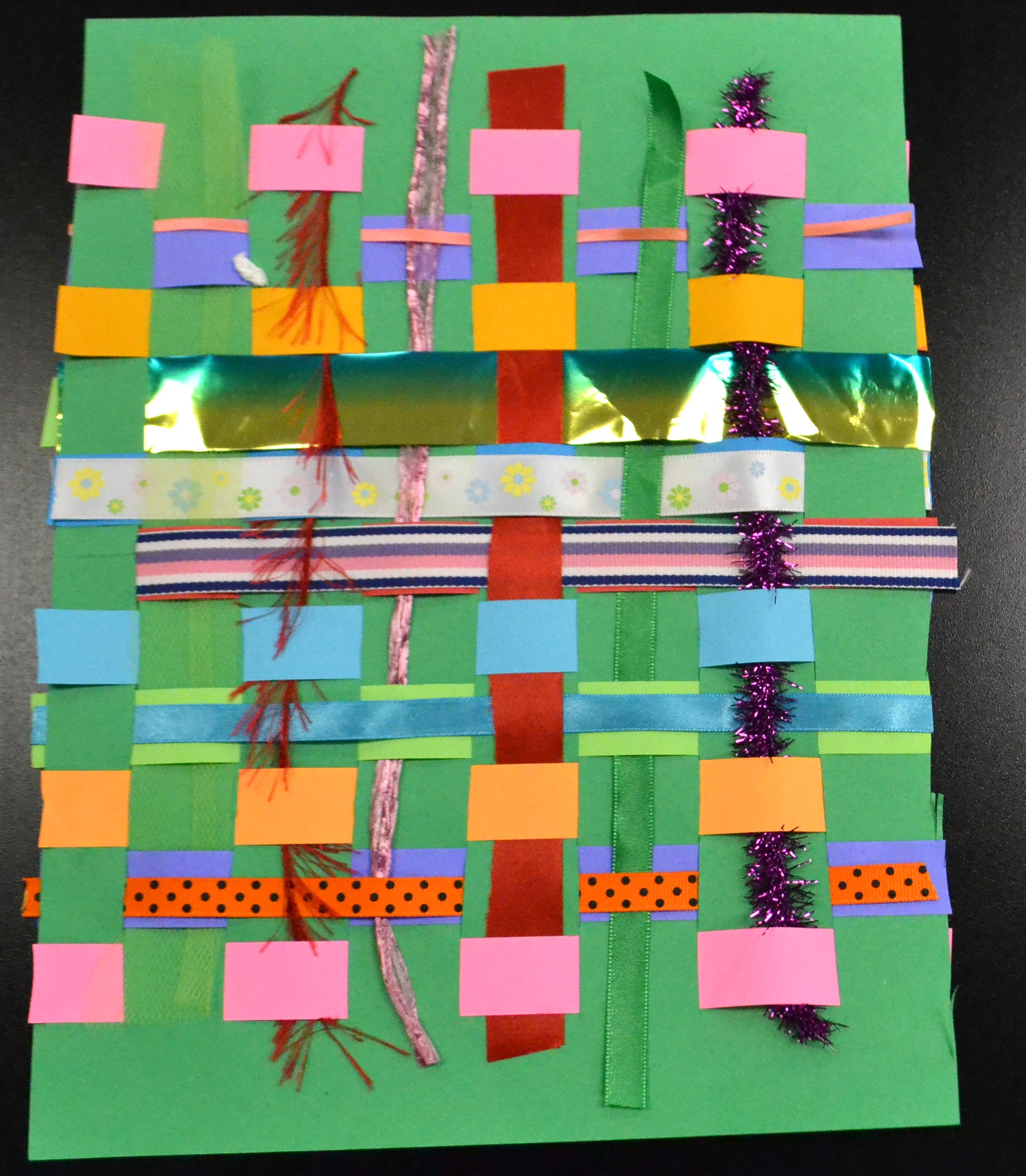 Paper strips, ribbon, pipe cleaners, and feathers woven through a green background