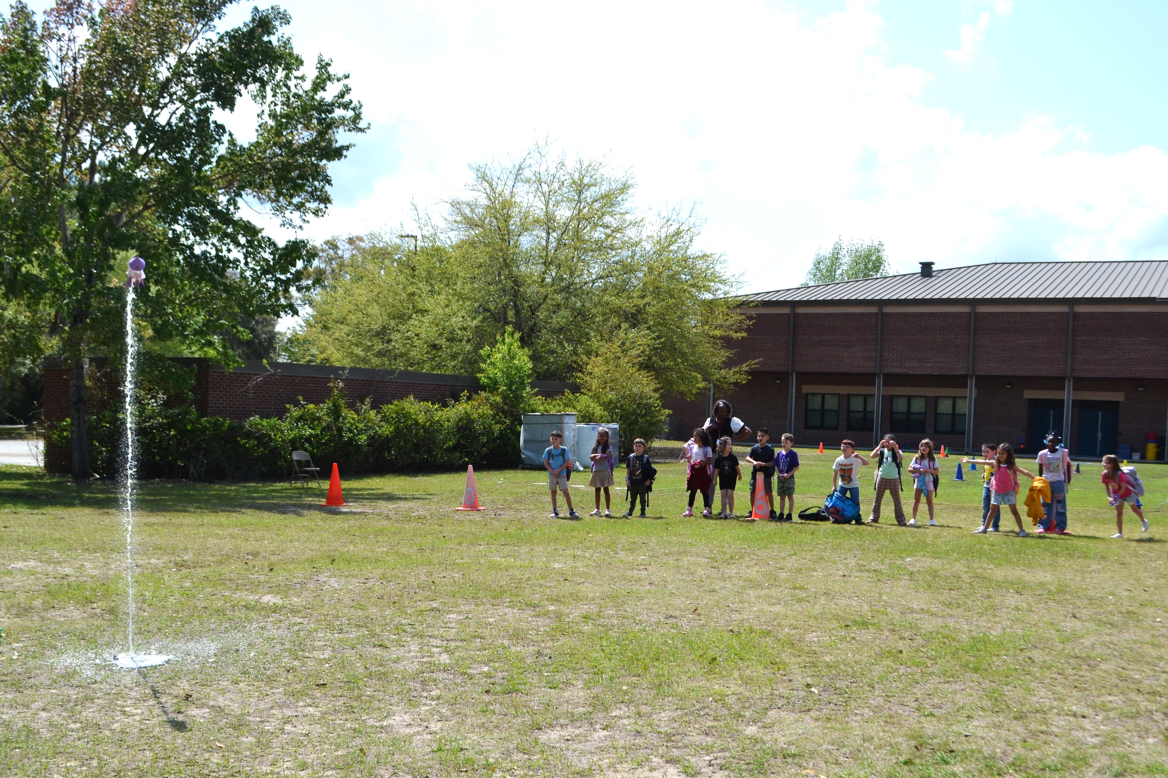 Students stand back as a soda bottle rocket launches into the air. A stream of white is seen beneath the rocket. 